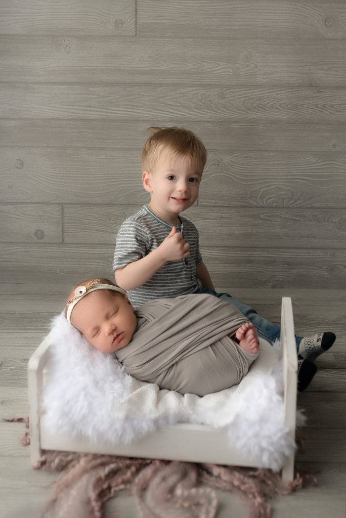 Proud big brother in sibling pose with new baby girl posed in prop in studio photography