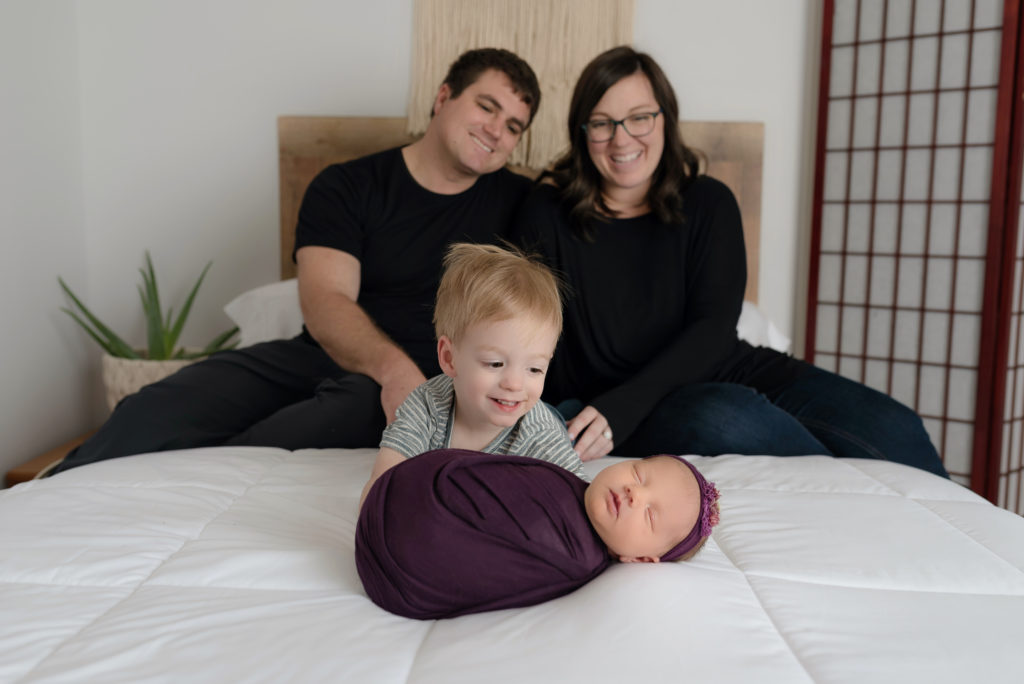 happy parents and toddler looking at newborn baby sister wrapped in purple on bed