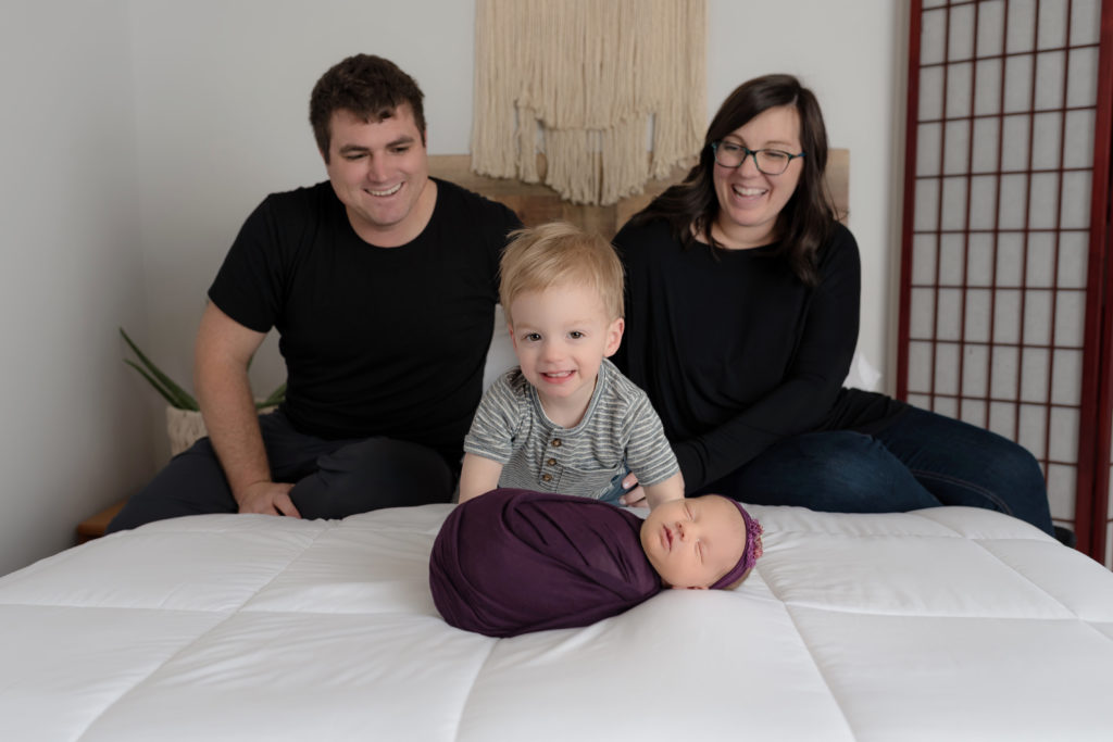 Smiling boy posed with baby girl with family on bed for newborn session