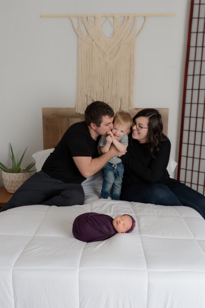 Family kiss from parents to son in background of baby girl posed on bed