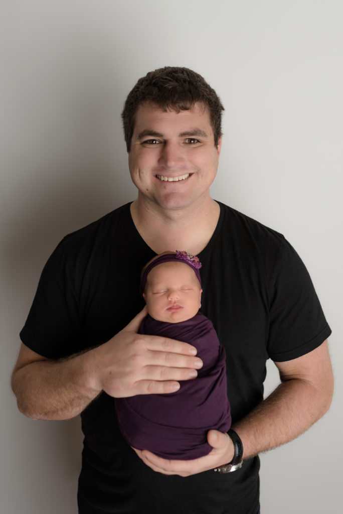 Dad smiling with newborn girl in hands facing out in charlotte studio
