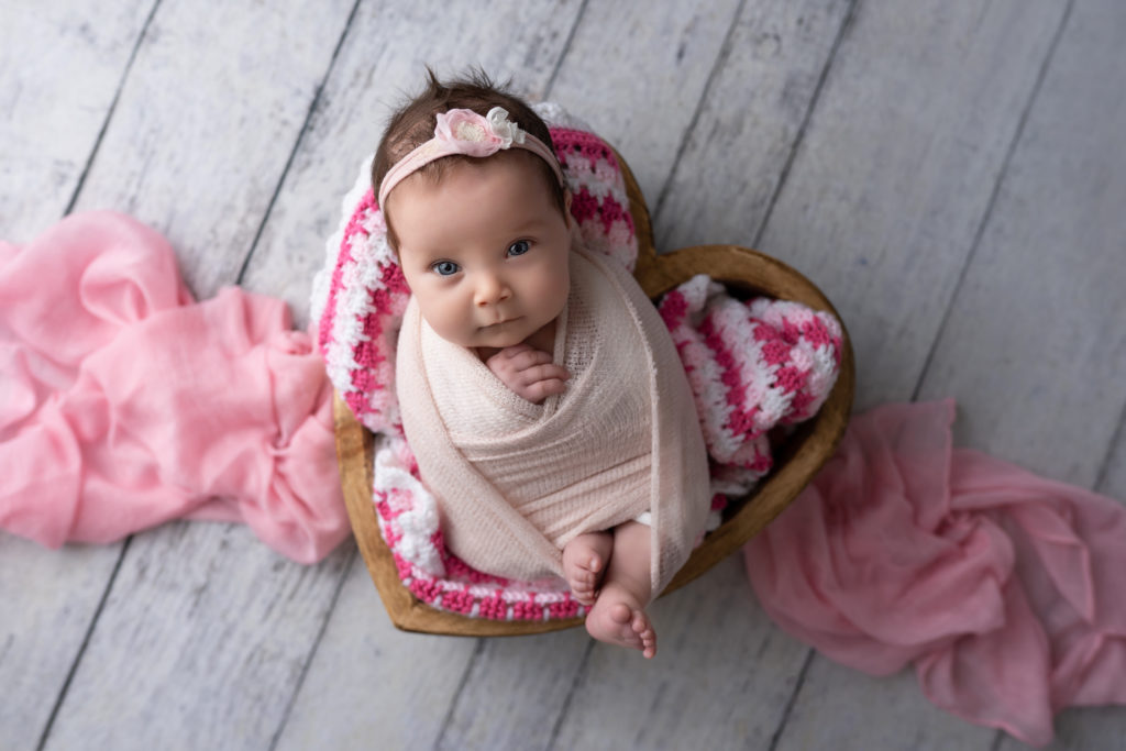 baby girl in pink in wooden heart bowl prop swaddled open eyes