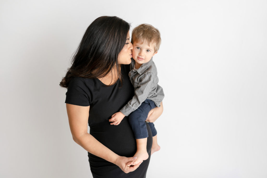 high end maternity photo mom kissing son on hip 
in studio on white
