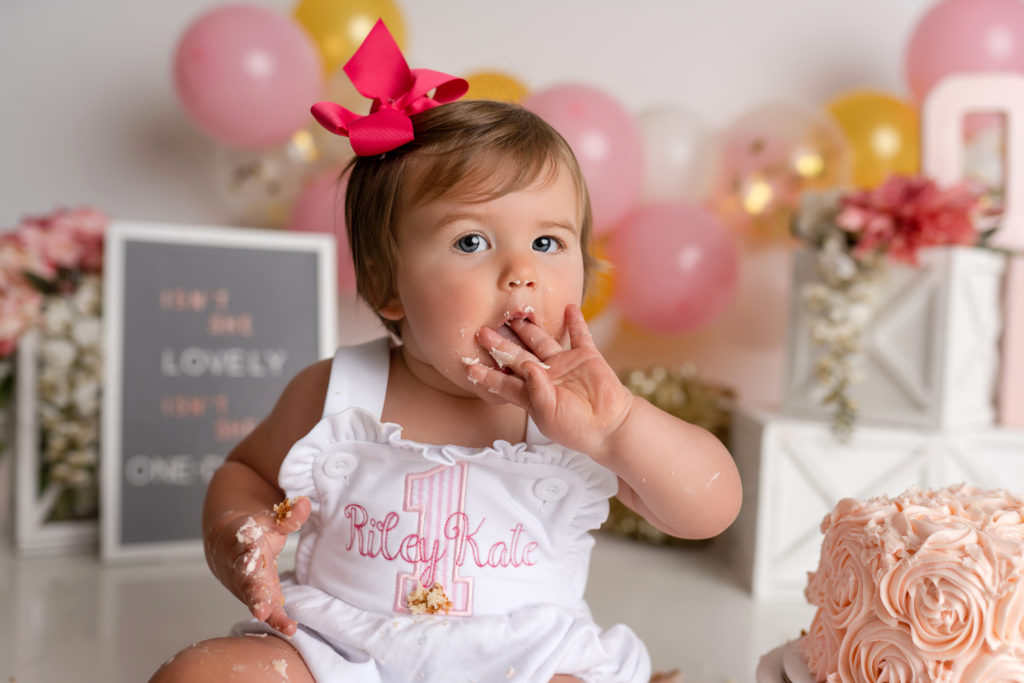 Little girl licking frosting off fingers in pink and gold theme cake smash in studio 