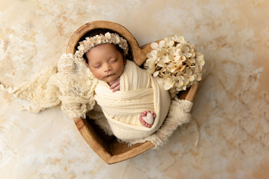 newborn girl wrapped in creamy yellow posed in heart shaped wooden bowl with flowers and heart between feet