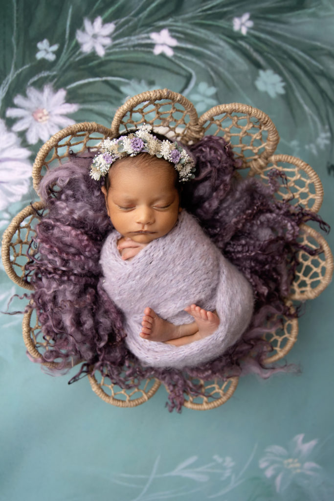 newborn girl wrapped in purple with floral headband posed in woven bowl
