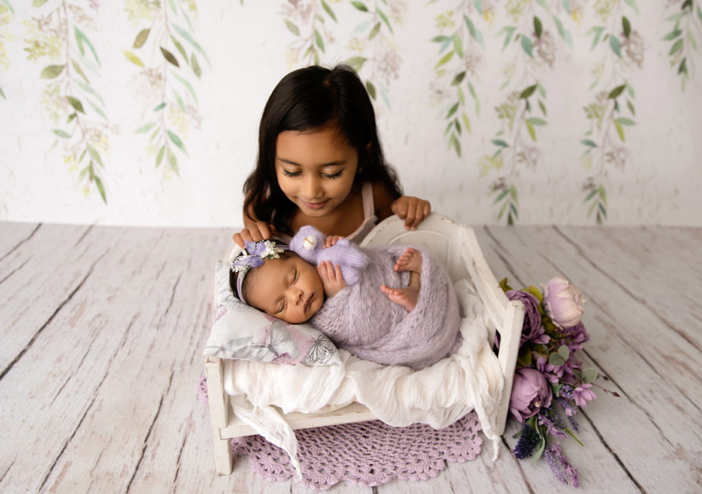 newborn girl wrapped in purple on bed prop with sibling