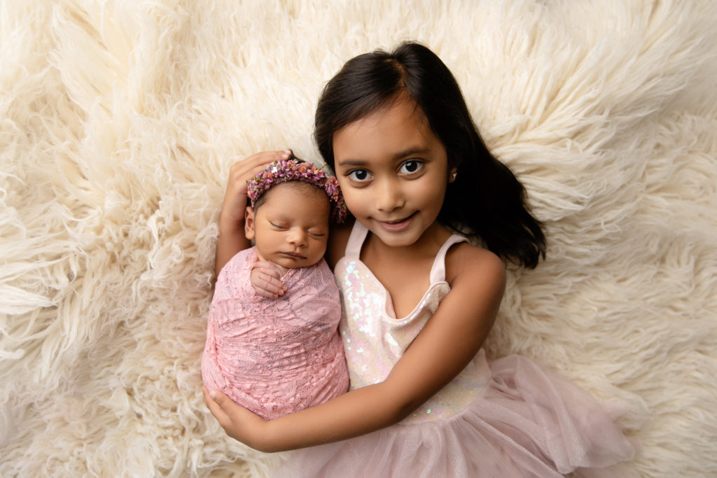 newborn girl wrapped in pink on cream fur with sister
