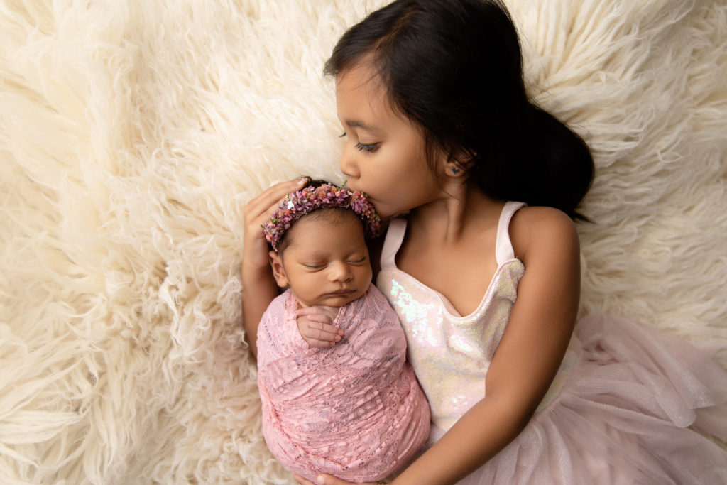 newborn girl wrapped in pink on cream fur with sister kissing head