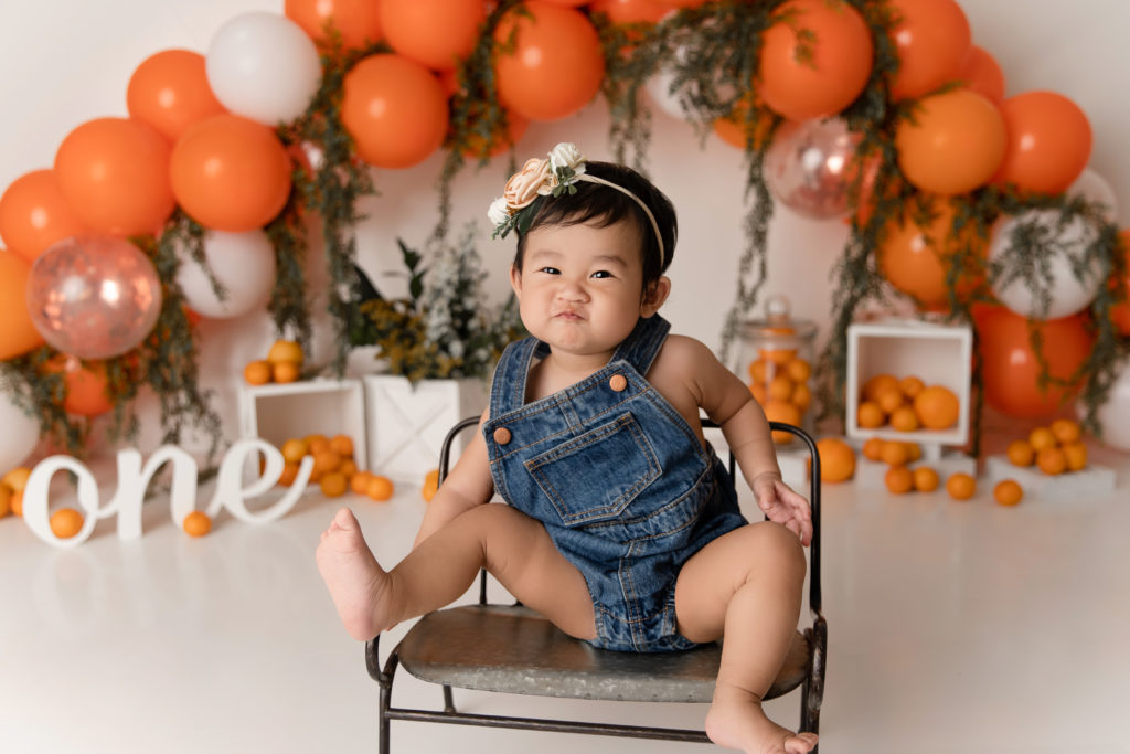 little girl in overalls on bench in front of orange cutie backdrop