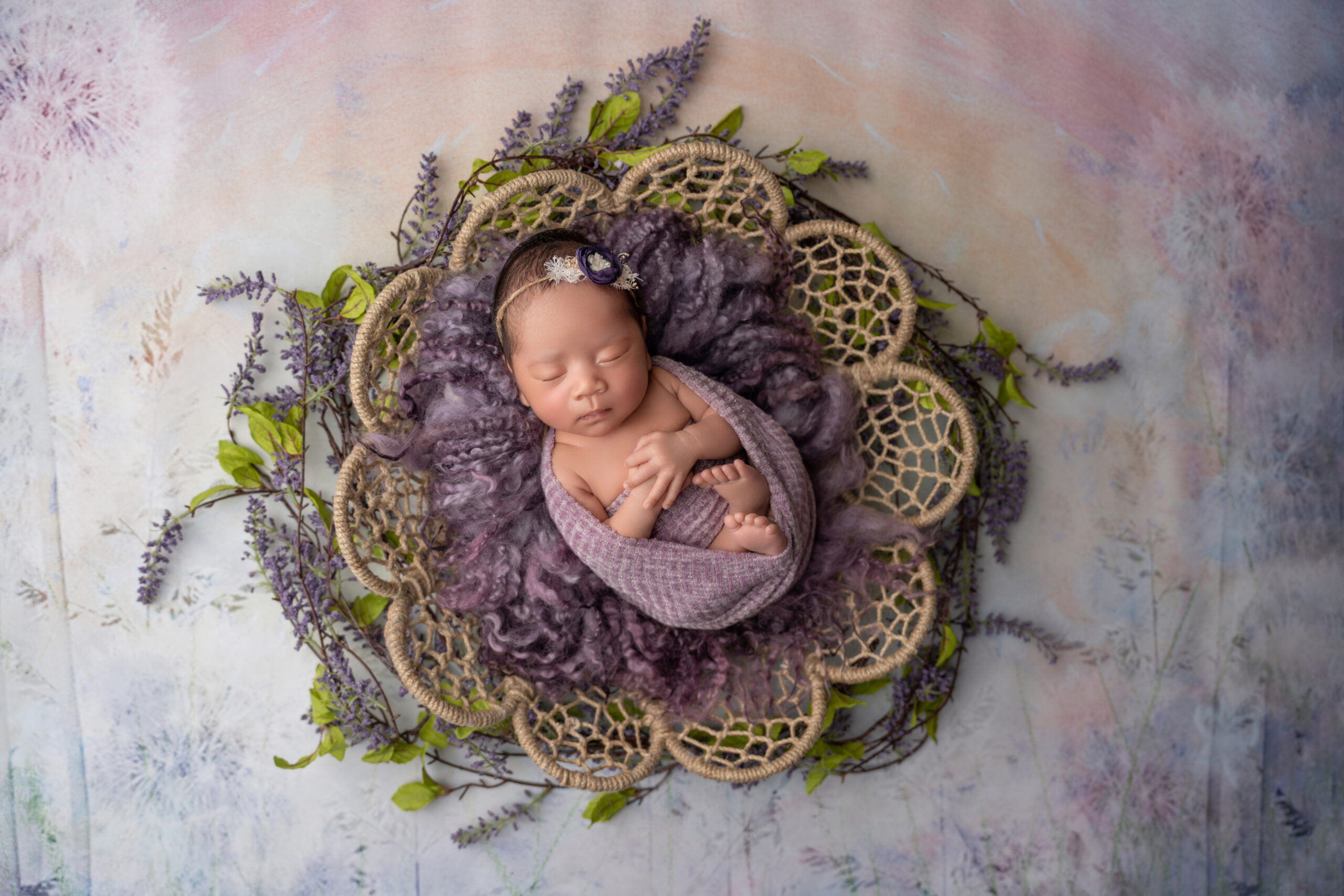 newborn girl wrapped in shades of purple with lavender under basket
